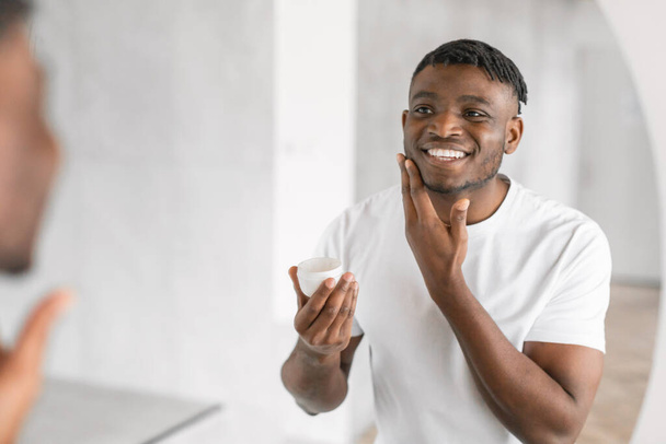 Happy young black man with a radiant smile stands in his bathroom, applying skincare product, holding moisturizer jar and touching soft facial skin near mirror indoor. Copy space - Zdjęcie, obraz