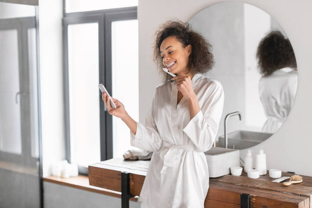 Atractiva dama negra vestida con albornoz de seda preparándose para cepillarse los dientes mientras usa el teléfono celular, se involucra en su rutina matutina en el baño interior. Tecnología, estilo de vida moderno - Foto, Imagen