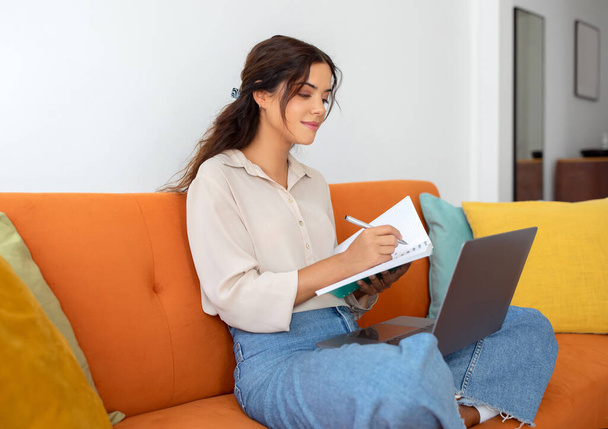Όμορφη νεαρή γυναίκα λαμβάνοντας σημειώσεις για να σημειώσετε, ενώ χρησιμοποιώντας το Laptop στο σπίτι, Ευτυχισμένη Millennial γυναικεία μελέτη με υπολογιστή στο εσωτερικό του σαλονιού, Απολαμβάνοντας εξ αποστάσεως εκπαίδευση, Ελεύθερος χώρος - Φωτογραφία, εικόνα