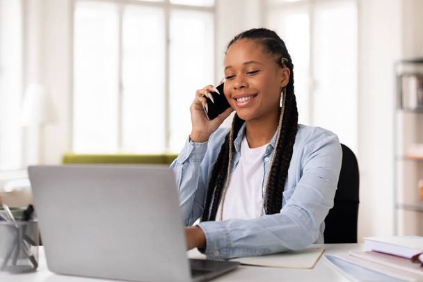 Radosna czarna nastolatka z plecionymi włosami angażująca się w miłą rozmowę na telefonie komórkowym podczas pracy na laptopie w jasnych warunkach domowych - Zdjęcie, obraz