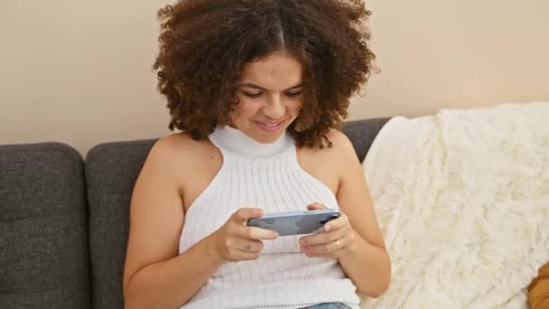 Eine junge Frau mit lockigem Haar genießt ihr Smartphone auf einer Couch im Haus und strahlt Freude und Entspannung aus. - Filmmaterial, Video