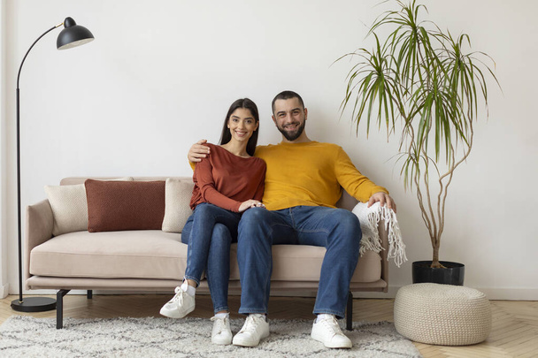 Happy Young Spouses Sitting On Sofa Embracing And Smiling To Camera, Romantic Caucasian Couple Posing At Home, Loving Husband Hugging His Wife. Love, Marriage And Romantic Relationship Concept - Photo, Image