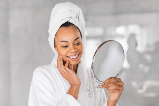 Μαύρη χιλιετή γυναίκα με μπουρνούζι που κρατάει στρογγυλό καθρέφτη, θαυμάζει την άψογη επιδερμίδα της στο μοντέρνο μπάνιο. Καθημερινή ρουτίνα περιποίησης και περιποίησης του δέρματος, επιδίωξη του τέλειου νεανικού δέρματος - Φωτογραφία, εικόνα