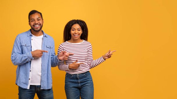 Nice Offer. Happy Black Couple Pointing Aside With Fingers, Indicating Copy Space, Young African American Man And Woman Showing Free Space For Your Advertisement, Standing On Yellow Background - 写真・画像