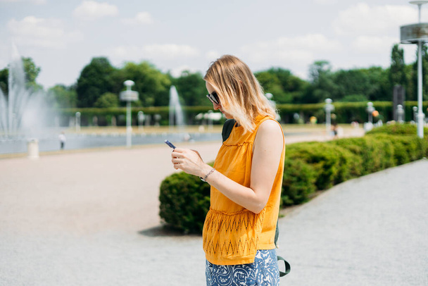 Sırt çantalı neşeli sarışın kadın şehir caddesinde yürürken akıllı telefonunu kontrol ediyor. Açık havada akıllı telefon kullanan 30 'lu yaşlardaki güzel bir kızın portresi. Kentsel yaşam tarzı konsepti. - Fotoğraf, Görsel