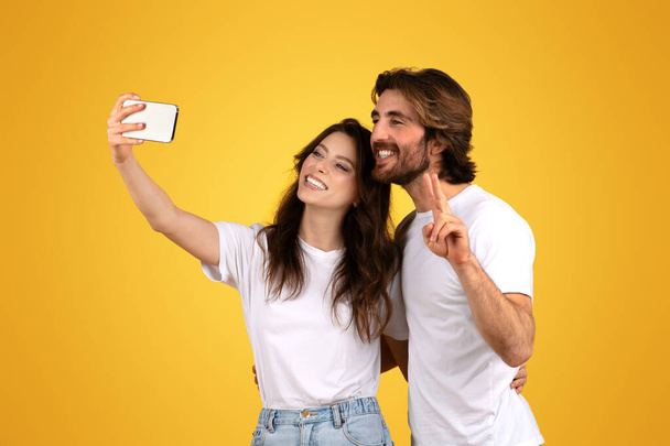 Joyeuse femme européenne et homme barbu prenant joyeusement un selfie, avec la femme tenant le téléphone et l'homme clignotant un signe de paix, tous deux en t-shirts blancs, sur un fond jaune - Photo, image