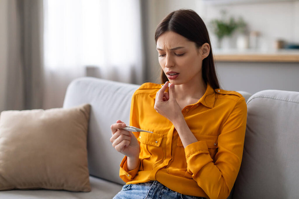 Mujer joven preocupada en blusa amarilla mirando el termómetro, sintiéndose mal, sentada en el sofá con expresión pensativa en el entorno del hogar - Foto, Imagen