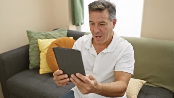 Volwassen man met behulp van tablet op comfortabele bank in een goed verlichte gezellige interieur - Video