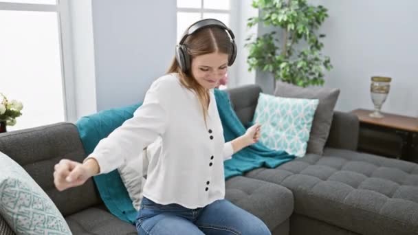 Een vrolijke jonge vrouw luistert naar muziek met een koptelefoon in een gezellige woonkamer, die het geluk uitdrukt. - Video