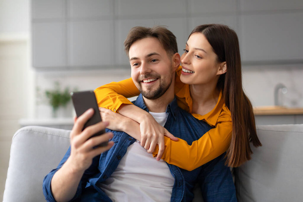 Улыбающиеся молодые супруги в повседневной одежде наслаждаются моментом вместе, снимая селфи по мобильному телефону, обнимаясь на удобном диване дома - Фото, изображение