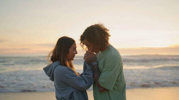 Молодые влюбленные встречаются на пляже Сансет. Нежный мужчина целует любящей женщине руки на вечернем побережье вблизи. Улыбающийся мужчина смотрит друг на друга с любовью. Два счастливых человека проводят время на природе - Фото, изображение