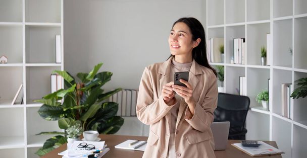 Азіатська бізнес-леді розмовляє по телефону на онлайн бізнес-зустрічі в сучасному домашньому офісі, прикрашеному пишними зеленими рослинами.. - Фото, зображення