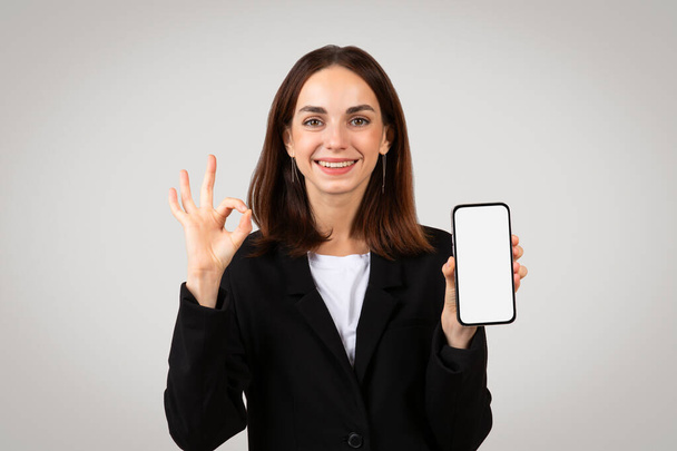 Mulher de negócios millennial caucasiana alegre mostrando uma tela de smartphone em branco e fazendo um sinal ok, indicando aprovação ou uma revisão positiva de um aplicativo móvel ou serviço - Foto, Imagem