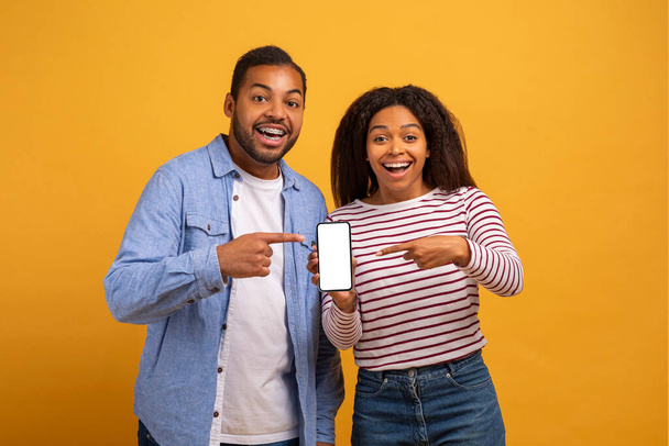 Geweldige app. Opgewonden zwarte man en vrouw wijzen naar lege smartphone met wit scherm in hun handen, Gelukkig jong Afro-Amerikaans paar aanbevelen van nieuwe mobiele applicatie, Mockup - Foto, afbeelding