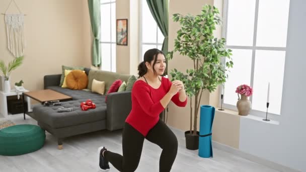 Jonge Spaanse vrouw die thuis traint in een woonkamer, lunges doet met een gefocuste houding. - Video