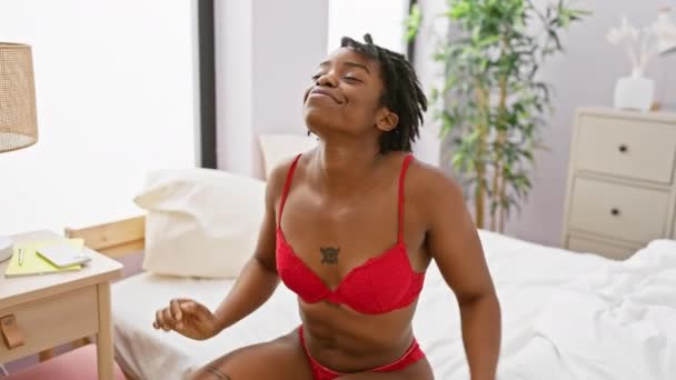 Μια Αφροαμερικάνα με κόκκινα εσώρουχα απλώνεται με χαρά στο φωτεινό της υπνοδωμάτιο. - Πλάνα, βίντεο