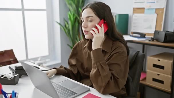 Keskittynyt nuori nainen osallistuu puhelinkeskusteluun työskennellessään kannettavan tietokoneen kanssa hyvin valaistussa toimistossa. - Materiaali, video