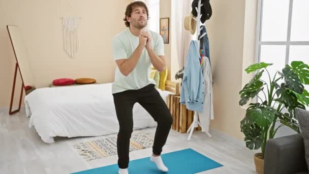 Mladý vousatý muž v neformálním oblečení cvičí jógu v úhledně organizované ložnici, vyzařuje klid a zdravý životní styl. - Záběry, video