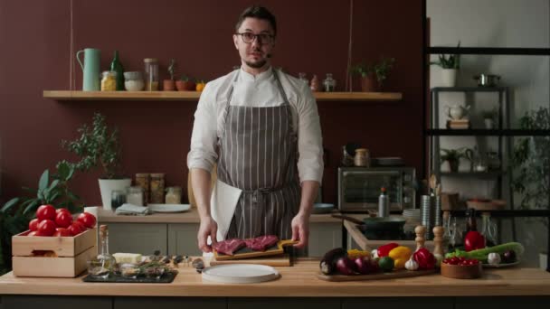 Medium of Caucasian male chef in uniform and eyeglasses speaking at camera and demonstrating two raw beef steaks on wooden cutting board while recording episode of cooking show in domestic kitchen - Footage, Video