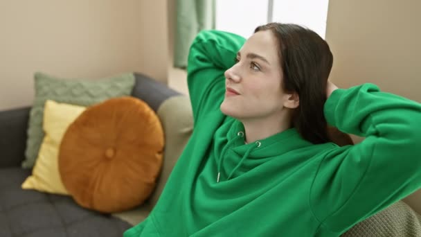 Relaxado jovem mulher em verde hoodie lounging em uma acolhedora sala de estar com almofadas coloridas. - Filmagem, Vídeo