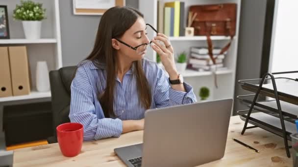 Egy gondolkodó fiatal spanyol nő az irodájában egy laptoppal, piros bögrével és stresszérzettel.. - Felvétel, videó