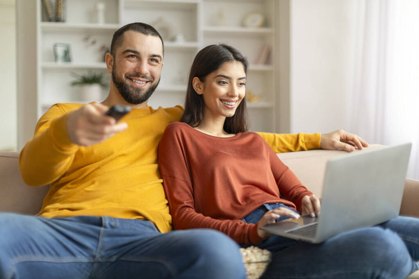 Счастливая любящая молодая пара проводит время вместе на диване дома, веселые кавказские супруги расслабляются в гостиной, улыбающийся мужчина смотрит телевизор и держит пульт дистанционного управления в то время как женщина с помощью ноутбука - Фото, изображение