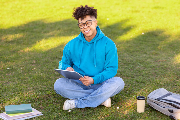 Sonriente estudiante brasileño navegando por Internet en tableta digital fuera, aprendiendo en el parque de la ciudad en el césped verde, estudiando con libros de ejercicios y mochila, mira a la cámara con sonrisa - Foto, Imagen