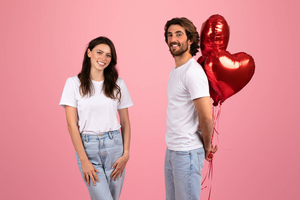 Smiling european woman and smiling man in white t-shirts, with the man holding a bunch of red heart-shaped balloons, celebrating a romantic occasion on a pink background, studio - Photo, Image