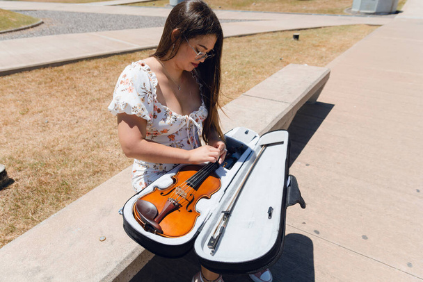 Latina busker γυναίκα κάθεται στο δρόμο βάζοντας βιολί στην περίπτωσή της, αφού κάνει τέχνη και παίζει βιολί. - Φωτογραφία, εικόνα