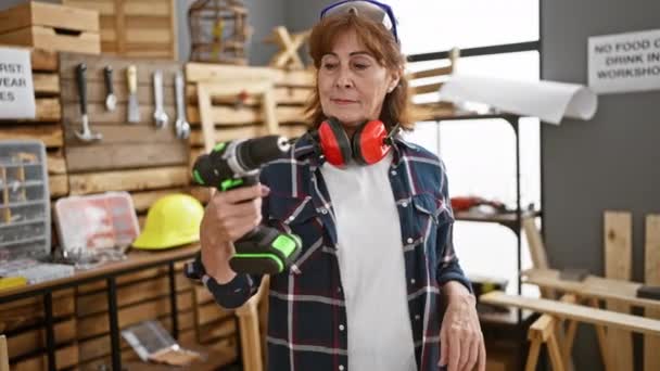 Kendinden emin orta yaşlı kadın marangoz, neşeli bir şekilde gülümseyen, el kol hareketi yapan dost canlısı marangozluk atölyesinde parmaklarıyla işaret eden, mükemmel matkap işi yapan - Video, Çekim