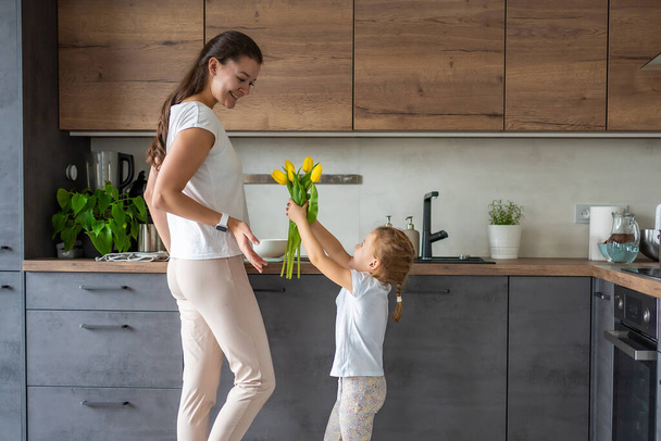 Little girl giving yellow tulips to her mum, while woman cooking on the kitchen. Mothers day, birthday or International Womens Day 8 march concept. High quality photo - Фото, изображение