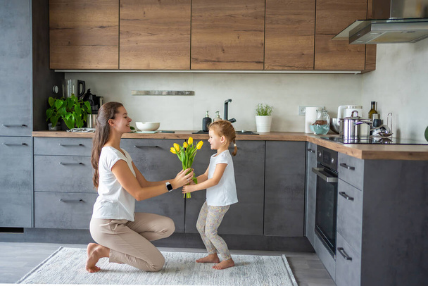 Маленька дівчинка дарує квіти своїй мамі, поки жінка готує на кухні. День матері, день народження або Міжнародний жіночий день 8 березня концепція. Високоякісна фотографія - Фото, зображення
