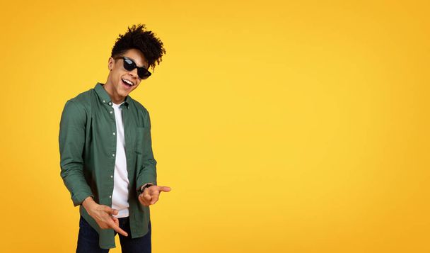 Stílusos millenniumi afro-amerikai srác göndör haj visel alkalmi ruházat és napszemüveg gesztikuláló sárga stúdió háttér, mutató kamera és mosolygós, panoráma fénymásolás tér - Fotó, kép