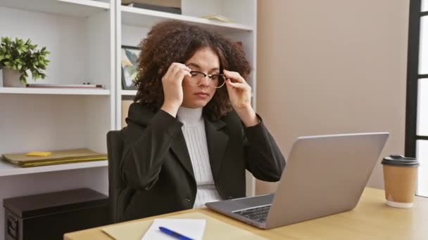 Una joven hispana con el pelo rizado examina el papeleo en el escritorio de su oficina, rodeada de computadora portátil y café. - Metraje, vídeo