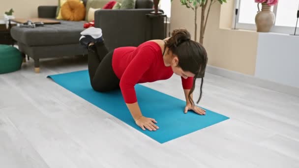 Donna ispanica che si allena in un moderno soggiorno su un tappeto yoga blu, indossando un top rosso e leggings neri. - Filmati, video