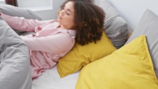 Молодая женщина с вьющимися волосами растягивается в своей спальне с розовой ночной рубашкой и желтыми подушками. - Кадры, видео