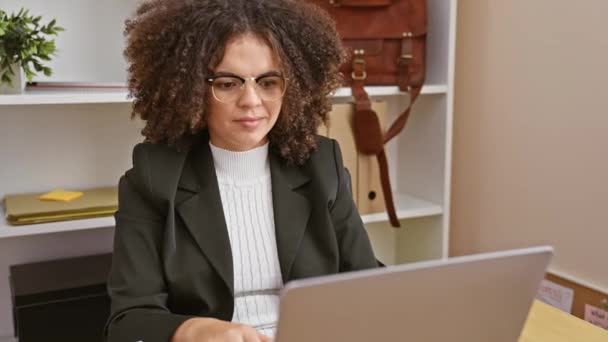 Mujer hispana con el pelo rizado sonriendo, utilizando el ordenador portátil en un entorno de oficina moderno - Imágenes, Vídeo