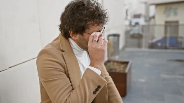 Een jonge Spaanse man met een baard verwijdert zijn bril, lijkt moe op een stedelijke straat achtergrond, portretteert een openhartig moment. - Video