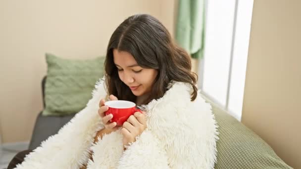Hispanic woman in a cozy living room smiles while holding a red cup, depicting a relaxing moment at home. - Footage, Video