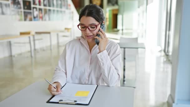 Una mujer hispana enfocada en múltiples tareas hablando por teléfono y escribiendo notas en su oficina moderna. - Metraje, vídeo