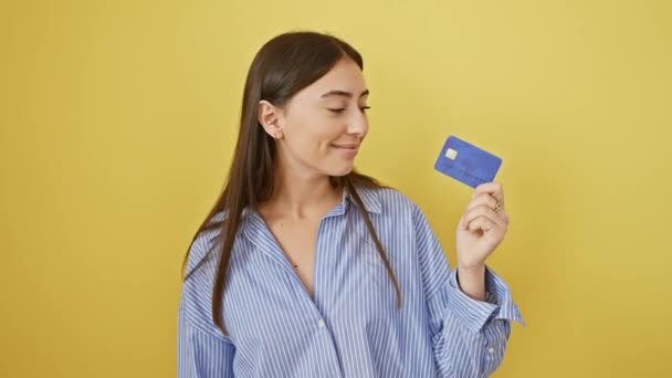 Радостная молодая латиноамериканка, мигающая пальцами, держа кредитную карту, излучает уверенность и счастье на изолированном желтом фоне. - Кадры, видео