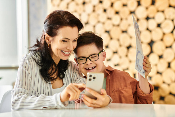 веселая женщина в уютной одежде со своим сыном с синдромом Дауна, смотрящая на меню на смартфоне - Фото, изображение