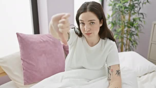 Mujer joven enojada sentada en la cama, mostrando aversión con los pulgares hacia abajo gesto en su pijama, retrato de mal humor en el interior - Imágenes, Vídeo