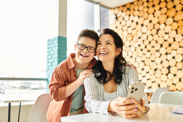 χαρούμενο αγόρι με σύνδρομο Down περνάει χρόνο με την όμορφη μητέρα του στο καφέ, κρατώντας smartphone - Φωτογραφία, εικόνα