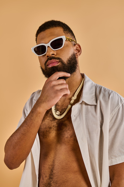Un hombre con barba llena lleva gafas de sol y un collar, exudando un comportamiento fresco y seguro de sí mismo mientras está de pie.. - Foto, Imagen