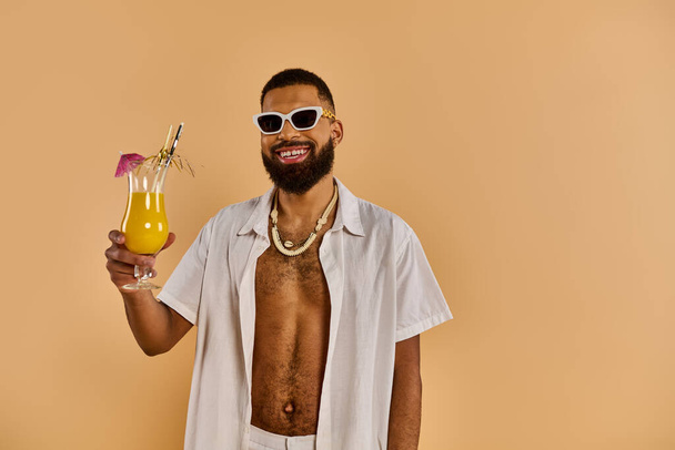Ένας κομψός άντρας με γυαλιά ηλίου κρατάει ένα ποτήρι χυμό, αποπνέοντας χαλάρωση και απόλαυση κάτω από τις ακτίνες του ήλιου. - Φωτογραφία, εικόνα