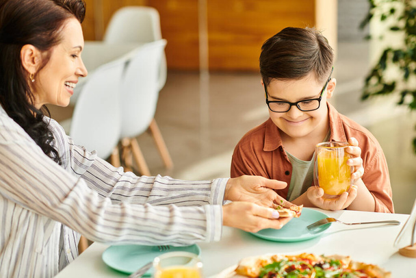 good looking mother eating pizza and drinking juice with her inclusive cute son with Down syndrome - Photo, Image