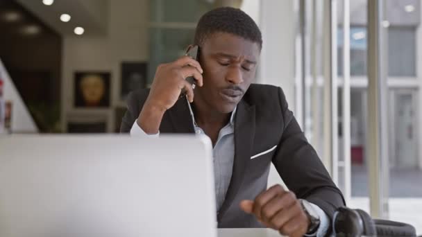 Hombre africano en traje multitarea con teléfono y computadora en el entorno de oficina. - Metraje, vídeo