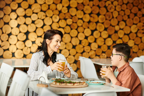gelukkig moeder eten pizza en drinken sap met haar inclusieve schattig zoon met Down syndroom - Foto, afbeelding