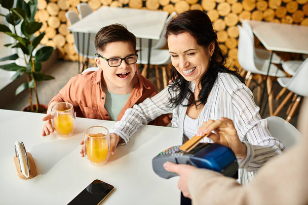 красивая веселая мать платит кредитной картой рядом с ее включительно сын с синдромом Дауна в кафе - Фото, изображение